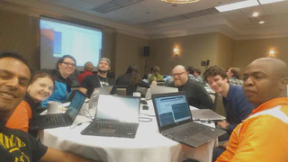 A selfie of 7 people sitting around a table with their laptops, smiling at the camera.